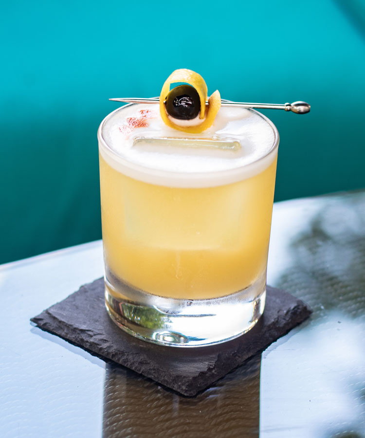 A golden elderflower whiskey sour cocktail in a rocks glass garnished with a lemon twist and cherry